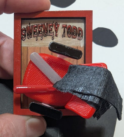 Small wooden frame with a swivel chair, knife and barber's cloth fabric, labeled 'Sweeney Todd', held by a hand.